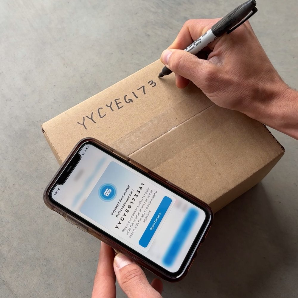 Hand writing the reference number on a parcel with a marker, phone beside it showing the number